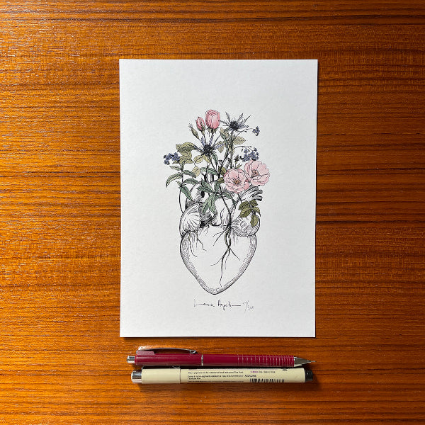 Ilustración de Laura Agustí de un corazón anatómico del que brotan rosas salvajes, cardos y nomeolvides en tamaño A5 sobre una mesa de madera