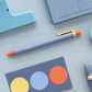 Bolígrafo de gel en azul lavanda, naranja y amarillo junto a  material de papelería azul