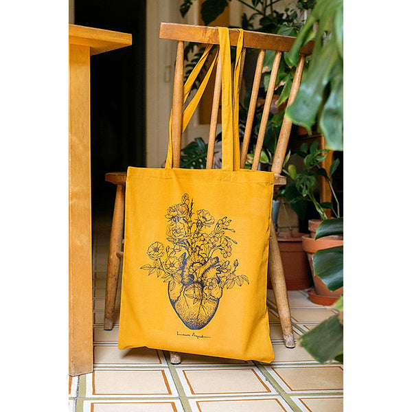 Bolsa de tela color cúrcuma con ilustración de un corazón anatómico del que brotan flores colgada de una silla en casa de Laura Agustí