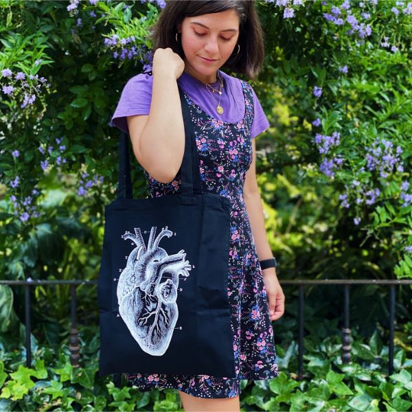 Mujer vestida con un estampado de flores y un bolso de tela negro con un corazón anatómico en un parque lleno de plantas verdes y flores moradas en Valencia