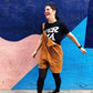 Mujer riéndose ante un muro de colores en Valencia con un peto de pana marrón y la camiseta de A fer la mà diseñada por la tienda de regalos Gnomo