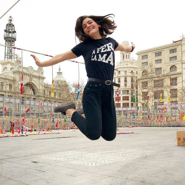 Mujer saltando en medio de la mascletá de la Plaza del Ayuntamiento de Valencia durante las Fallas con una camiseta negra en la que se lee la expresión típica "A fer la mà"