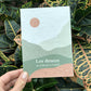 Tarjeta con semillas de Sheedo con un paisaje en tonos verdes y tierra con un sol naranja y el mensaje "Los deseos que se plantan se cumplen" sobre hojas de plantas