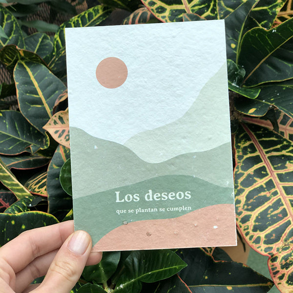 Tarjeta con semillas de Sheedo con un paisaje en tonos verdes y tierra con un sol naranja y el mensaje "Los deseos que se plantan se cumplen" sobre hojas de plantas