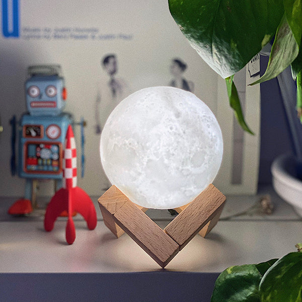 Lámpara con forma de luna pequeña sobre una estantería acompañada de un robot de juguete y un cohete