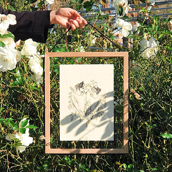 Mano sujetando um marco de madera y cristal con una ilustración de Laura Agustí sobre un fondo de plantas