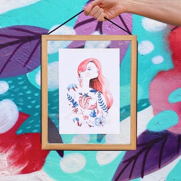 Mano sujetando un marco de madera y cristal con una ilustración de una mujer con flores y un corazón anatómico sobre fondo de colores