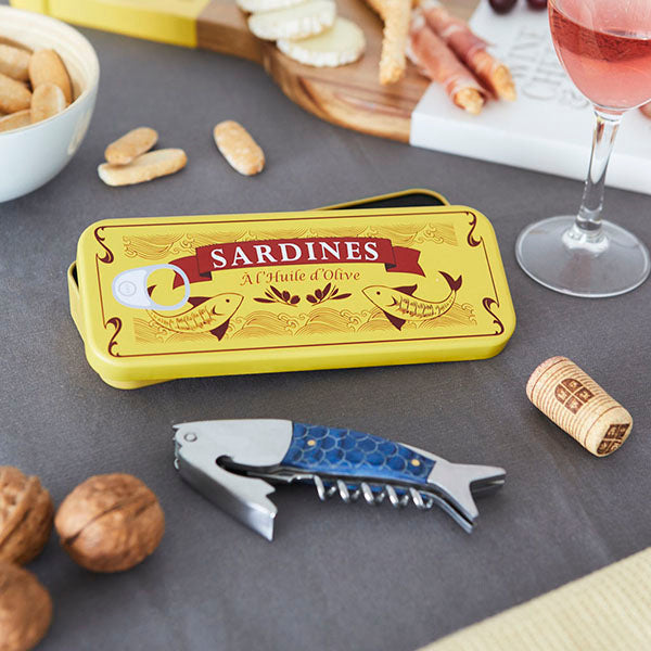Sacacorchos azul y metálico con forma de sardina dentro de una caja amarilla de lata en la que pone Sardines á l'huile d'olive rodeado de una copa de vino rosado y frutos secos