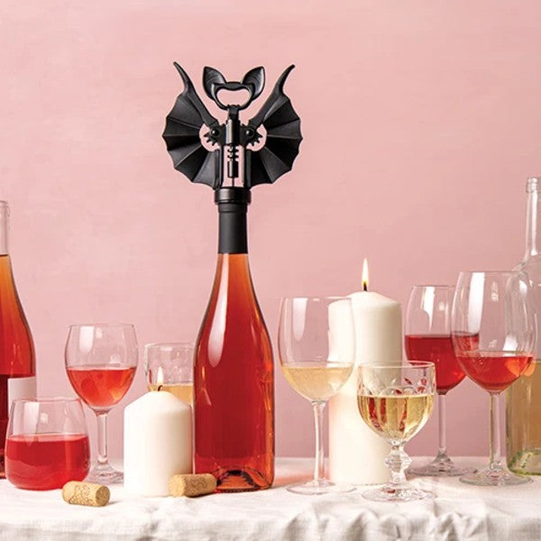 Sacacorchos de Ototo Vino con forma de murciélago negro con alas y colmillitos para usar como abridor abriendo una botella de vino rosado en una mesa puesta con velas y sobre un fondo rosa