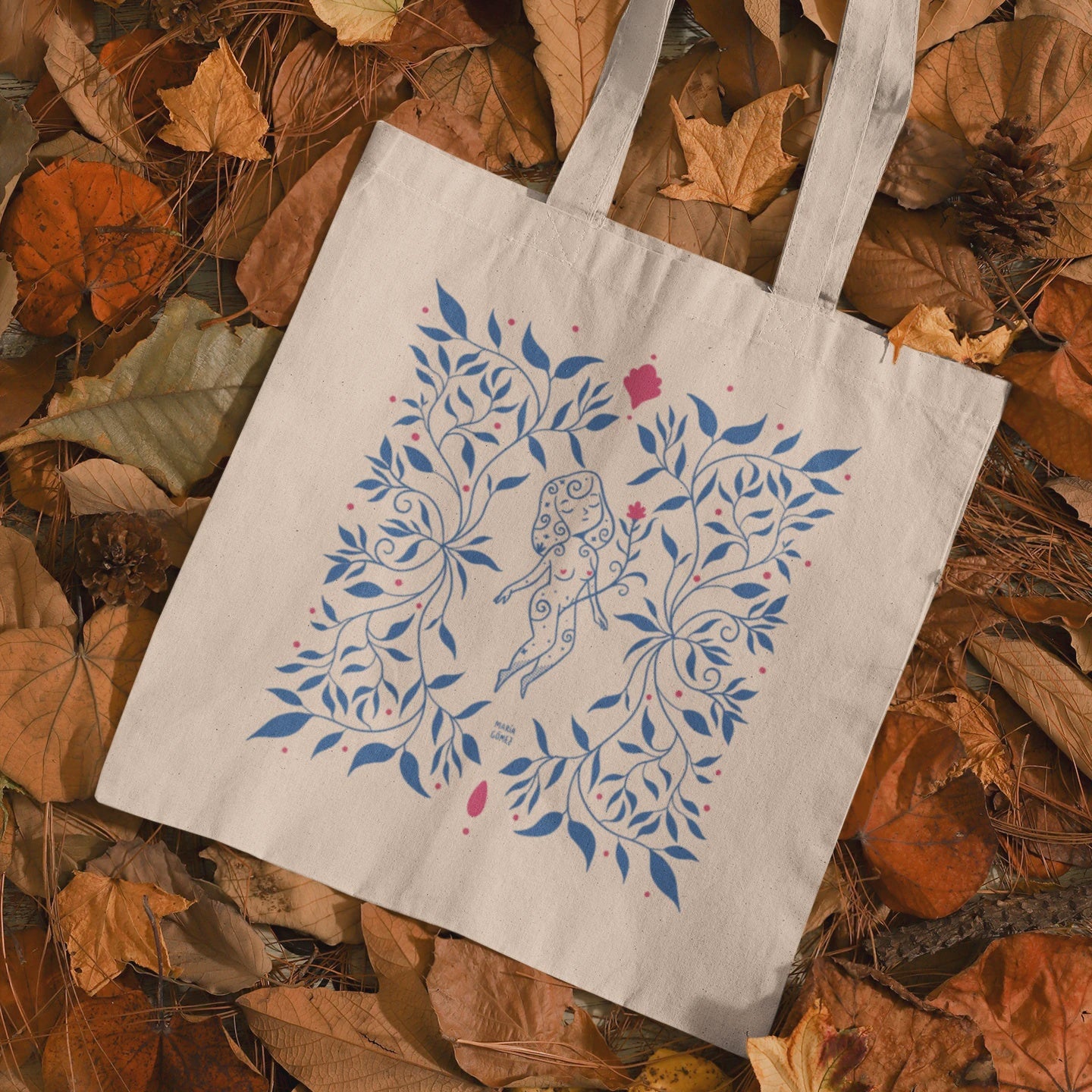 Bolsa de tela con una mujer flotando entre flores azules de María Gómez sobre un lecho de hojas secas