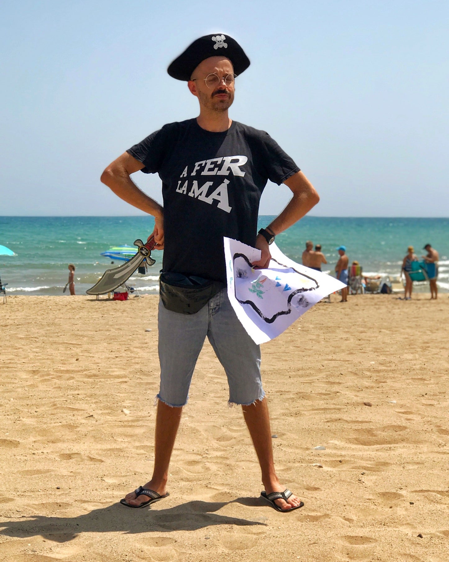 Chico con camiseta negra con inscripción A fer la mà, gorro y sable de pirata y un mapa del tesoro en la playa