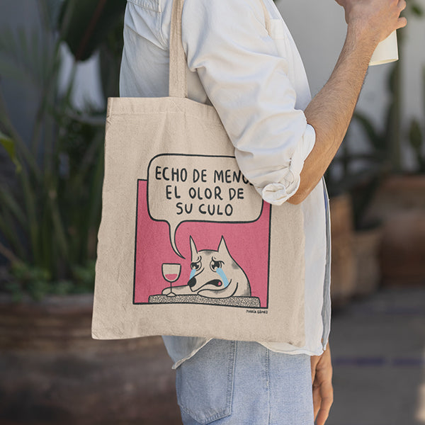Persona de perfil con la bolsa de tela ilustrada por María Gómez con un perro llorando con una copa de vino mientras dice "Echo de menos el olor de su culo