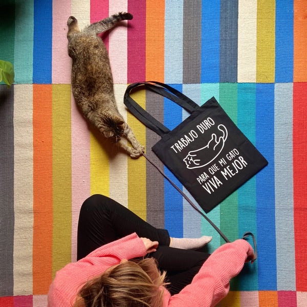 Una gata jugando con su dueña sobre una alfombra de colores y la bolsa negra de Trabajo duro para que mi gato viva mejor