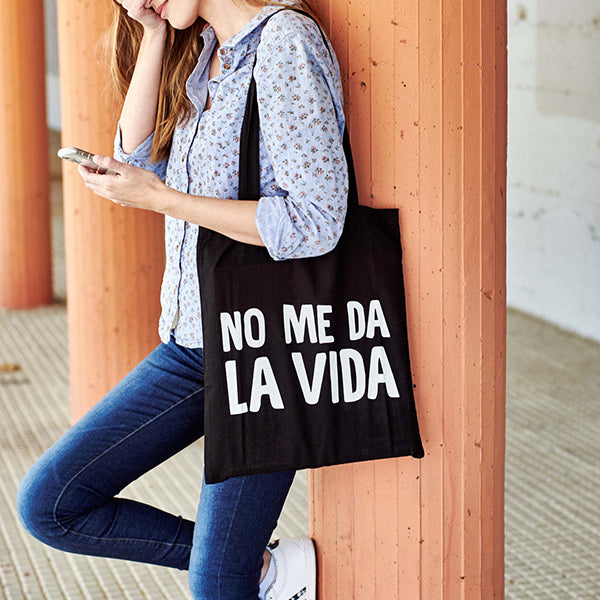 Mujer joven mirando el móvil con una bolsa de tela negra con la frase: "No me da la vida"